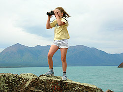 Photo courtesy of Bill and Anita Miller. A girl stands on a peak with binoculars with aqua ocean inlet below her.