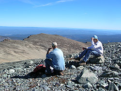 Photo courtesy of Bill and Anita Miller. Two people have lunch atop a peak that looks over an unending view of blue mountains.