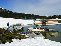 A plane parked near snowy shore with aluminum boat and people.