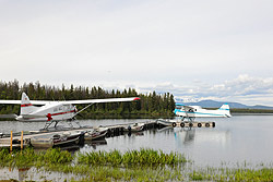 A red and white plane and a light blue and white plane are moored at a dock with mountain behind.