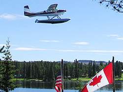 A maroon and white plane flies past an American and a Canadian flag on poles.