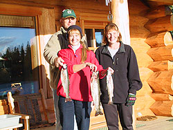 Three people standing on the deck of the lodge hold up Rainbow Trout.