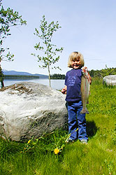 A little girl holds up a fish standing next to a rock.
