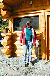 A man holds a large fish in front of log building.