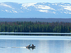 Two people going out fishing on the lake in a boat with snow on the mountains.